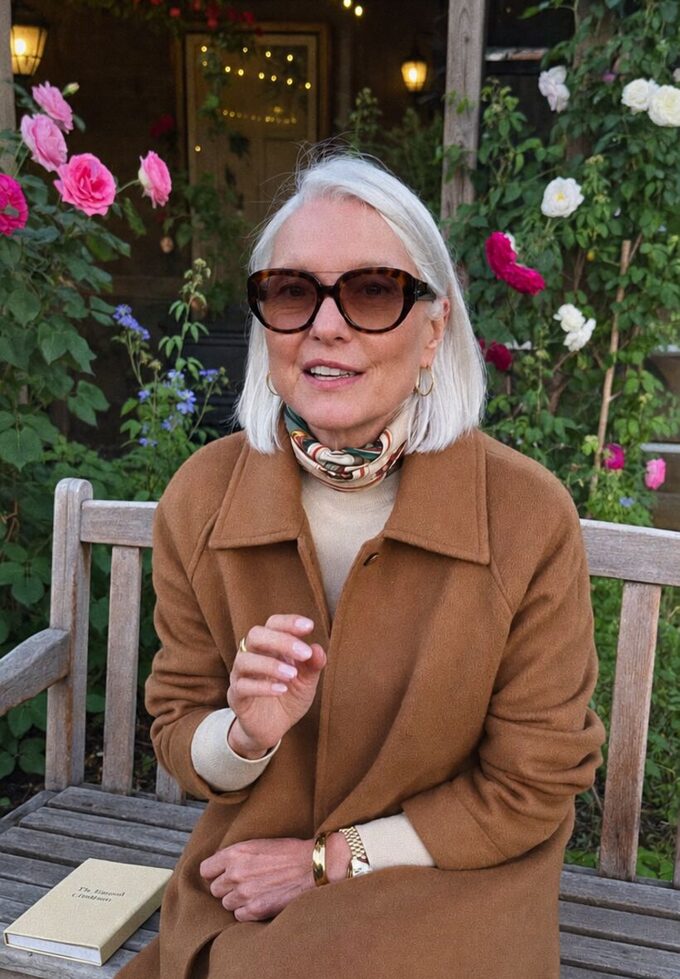 Eleanor, a woman in her sixties with a sleek white bob, tinted tortoiseshell sunglasses, a camel coat and a silk scarf, sitting on a garden bench among roses